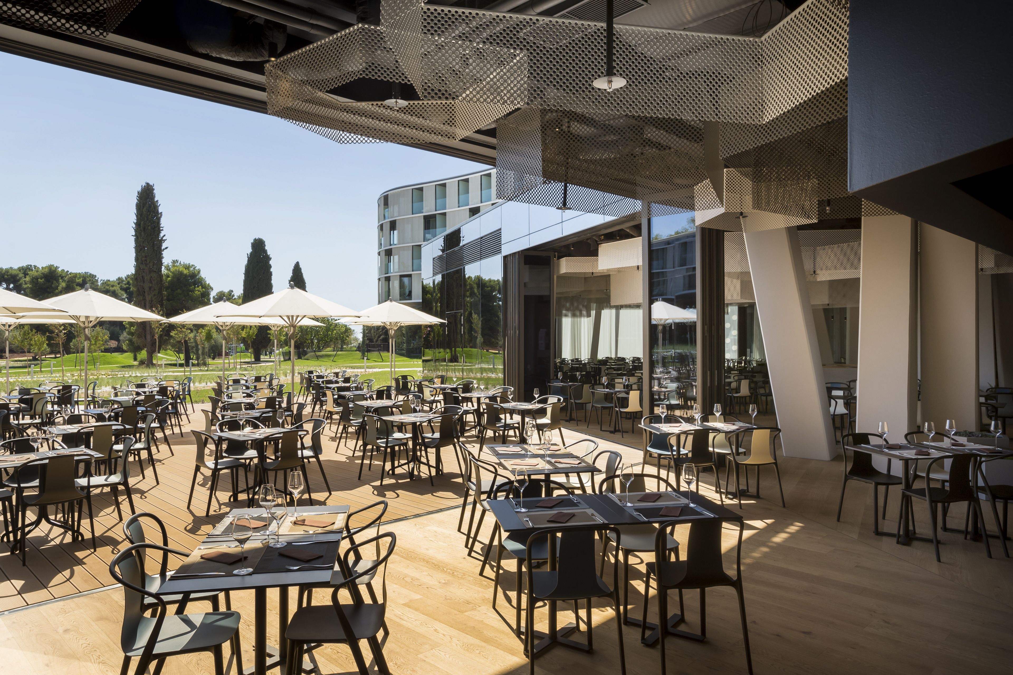 Un espacio de comedor al aire libre con mesas y sillas, grandes sombrillas y vistas a un edificio y árboles.