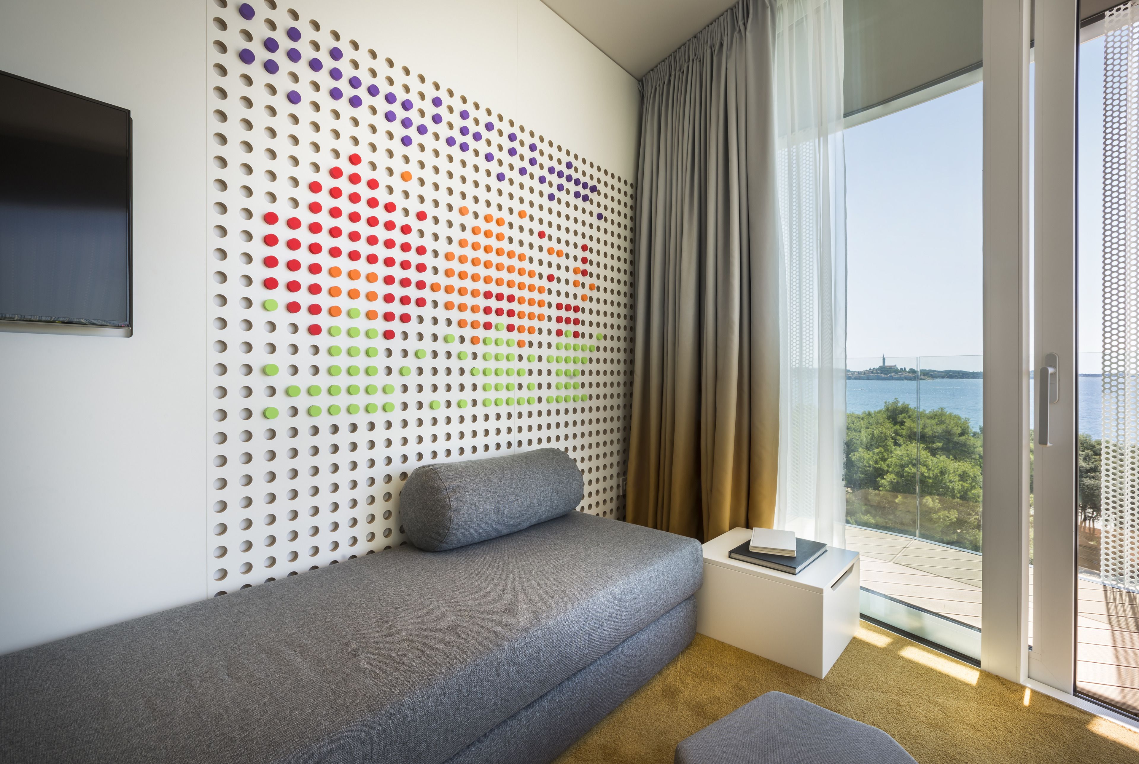 A modern hotel room with a sofa, a wall with a colorful dot pattern, a TV, and a view of the sea.
