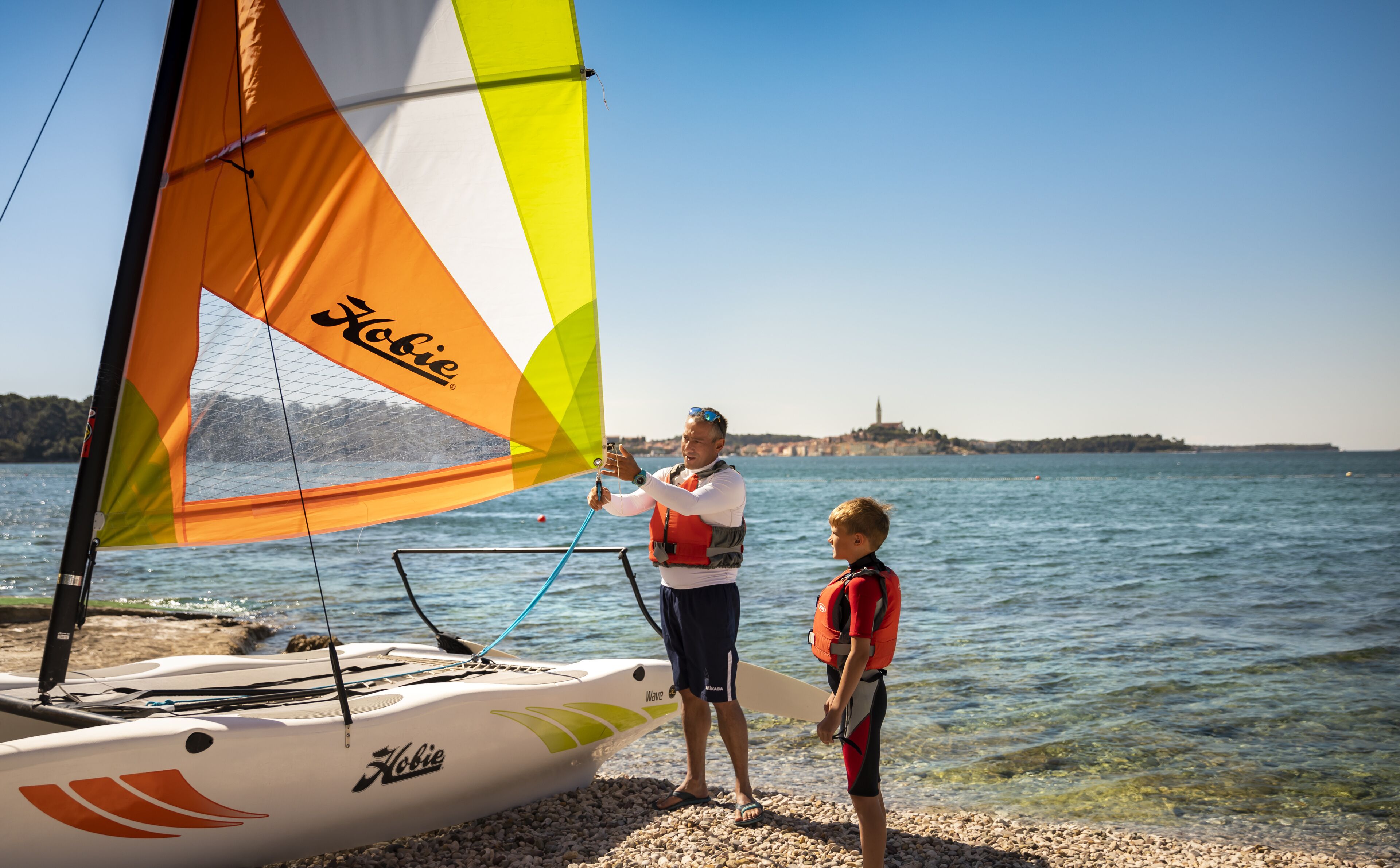 En segelbåt med ett färgglatt segel och namnet ”Hobie” på skrovet låg förtöjd vid en strand med småsten, där vattnet var klart och kustlinjen låg en bit bort.