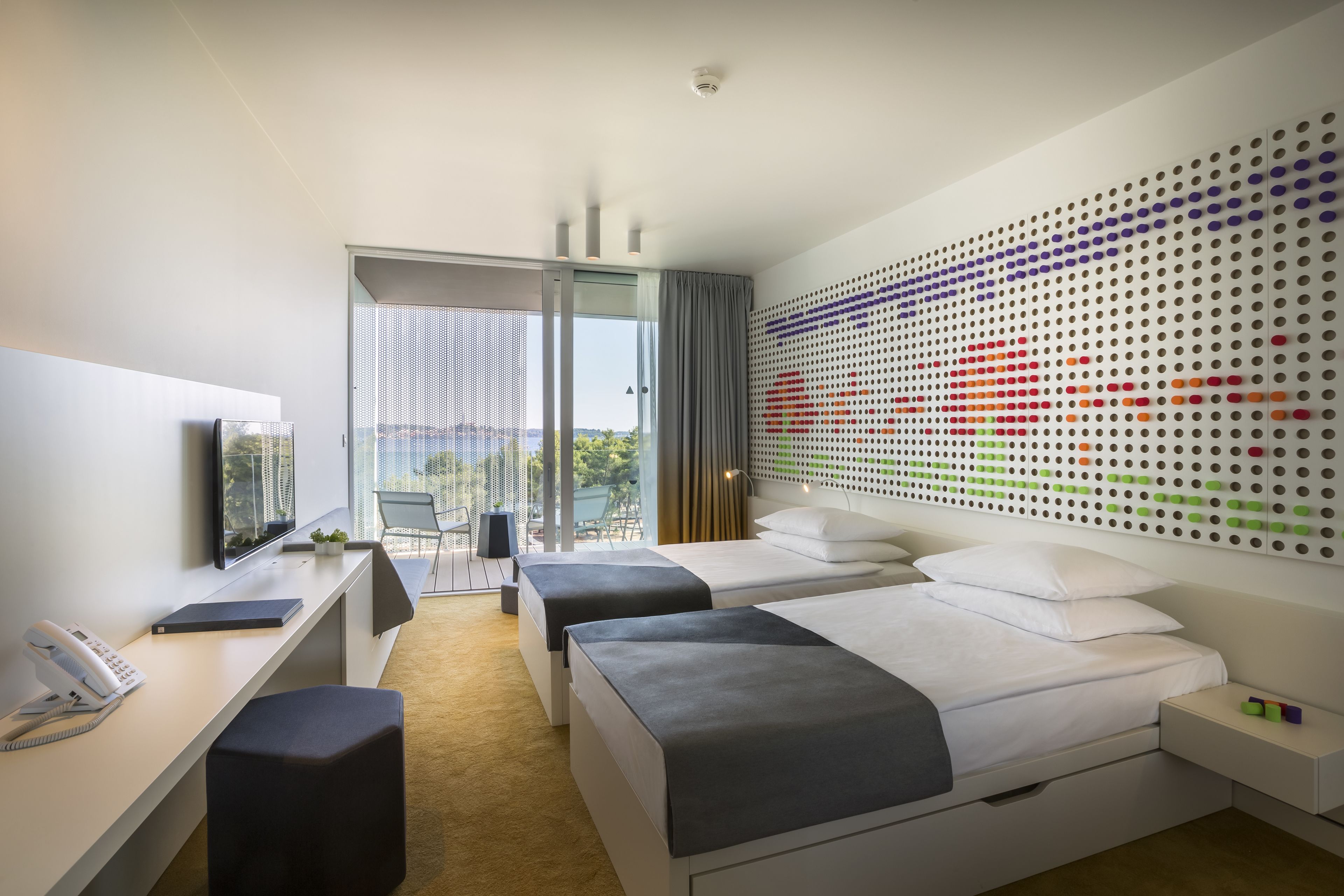 A modern hotel room with a desk, two beds, a large window, and a colorful dot-patterned wall.