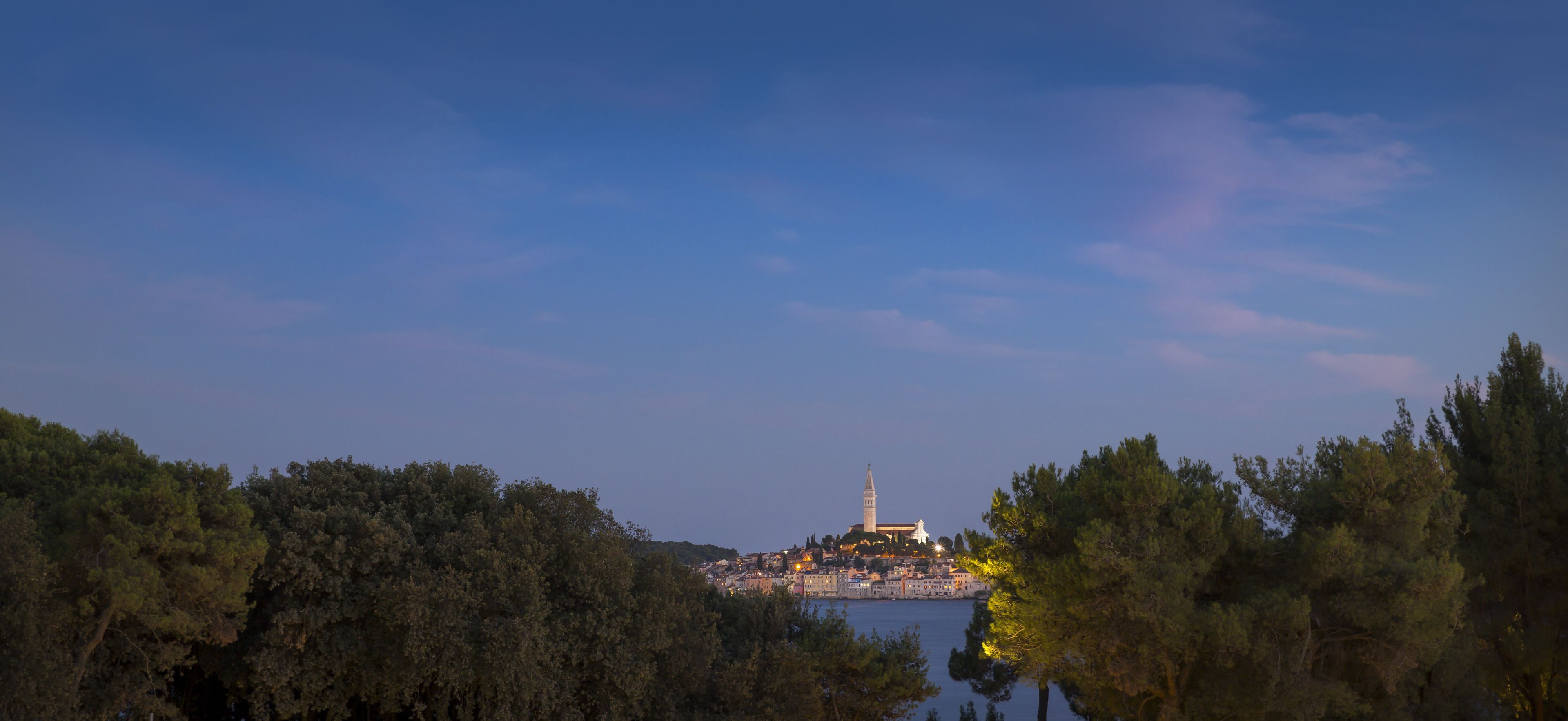 A coastal cityscape with a prominent tower and a cathedral, surrounded by dense green trees.