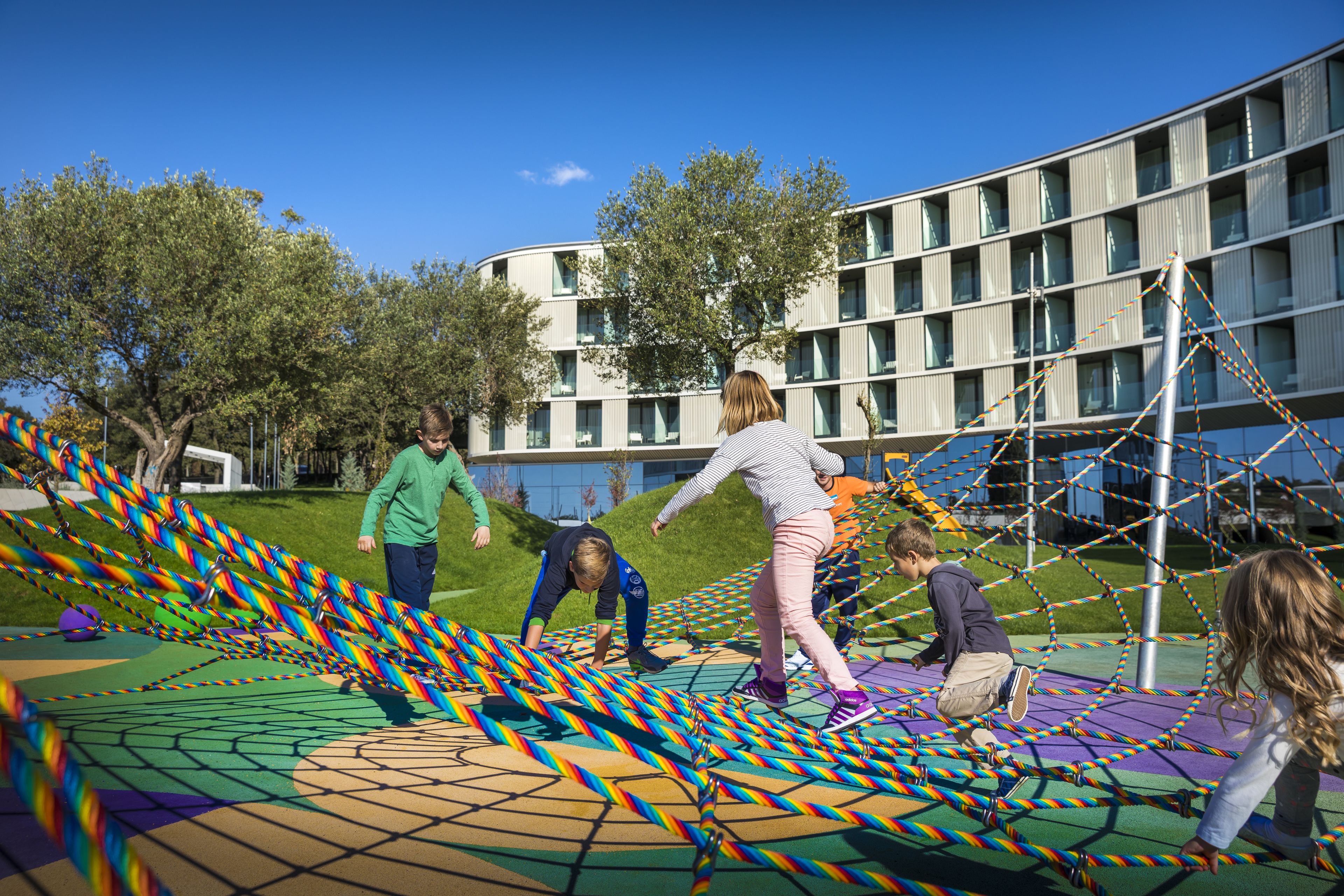Children playing on a colorful rope bridge in an outdoor area with a modern building in the background.