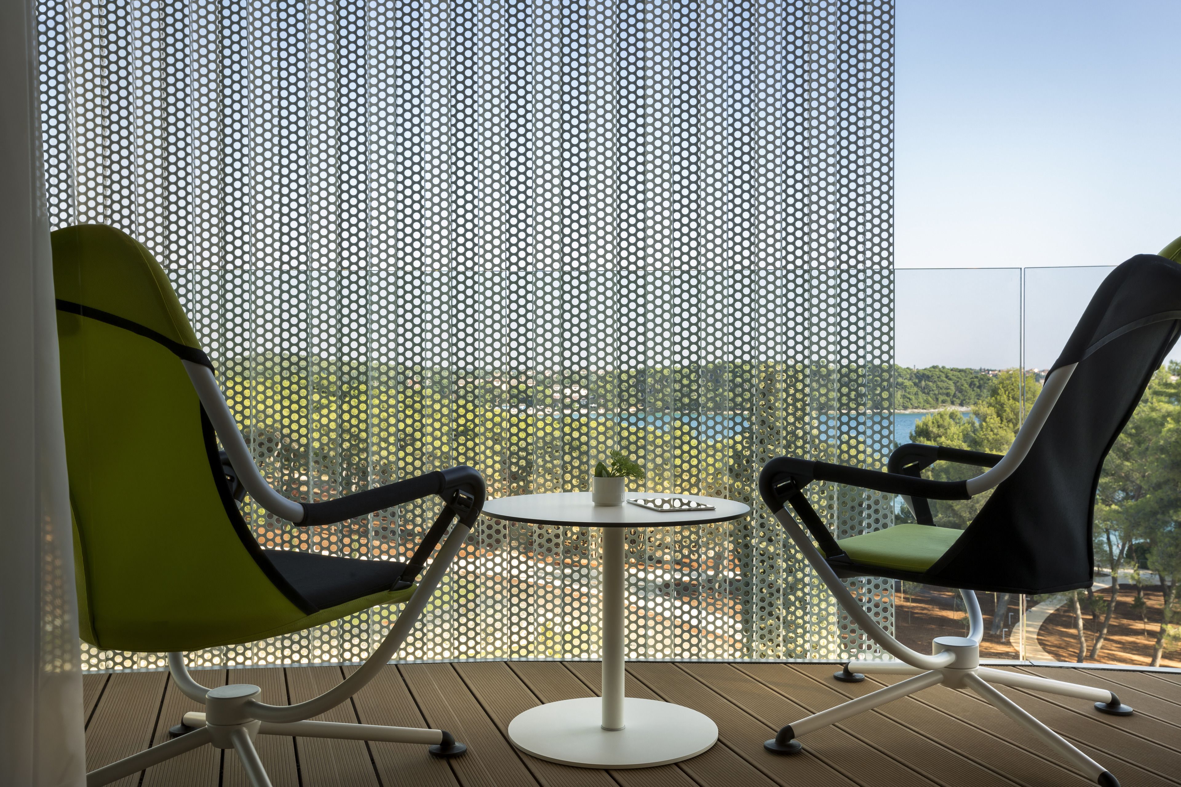 A balcony with two chairs and a small table, a perforated screen, and a view of greenery and buildings.