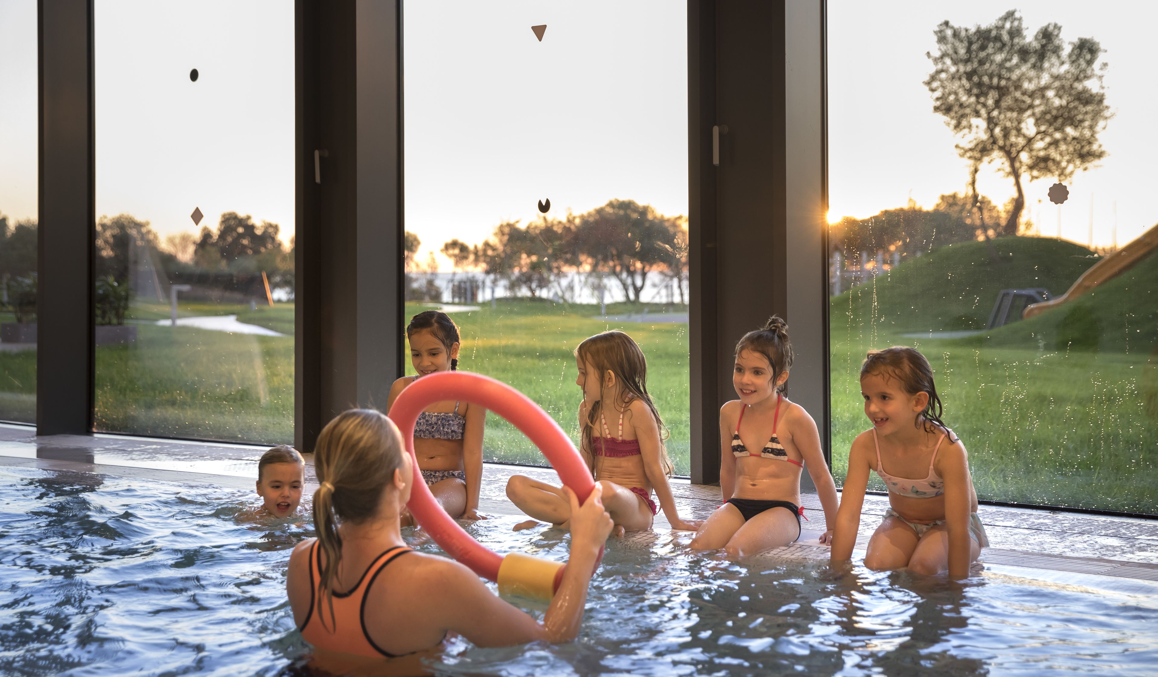 Une piscine avec des parois en verre transparent, donnant sur un parc, et un coucher de soleil.