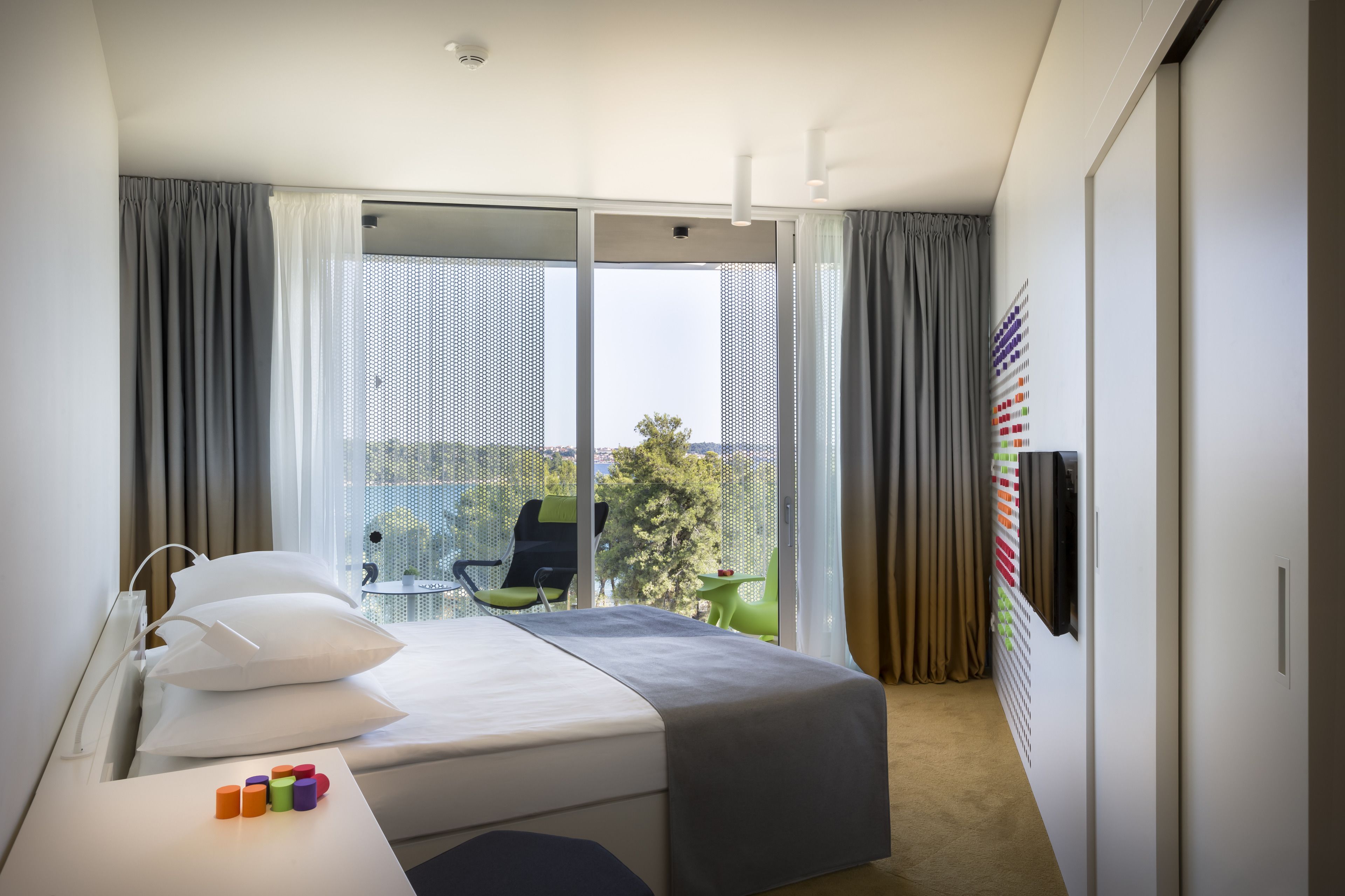 A modern hotel room with a large bed, a desk with colorful blocks, a chair, and a view of the outdoors through large windows.