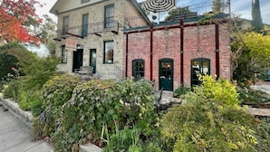 Un bâtiment à deux étages, doté d’une façade en pierre et d’une partie en briques rouges, avec un balcon et un petit espace jardin.
