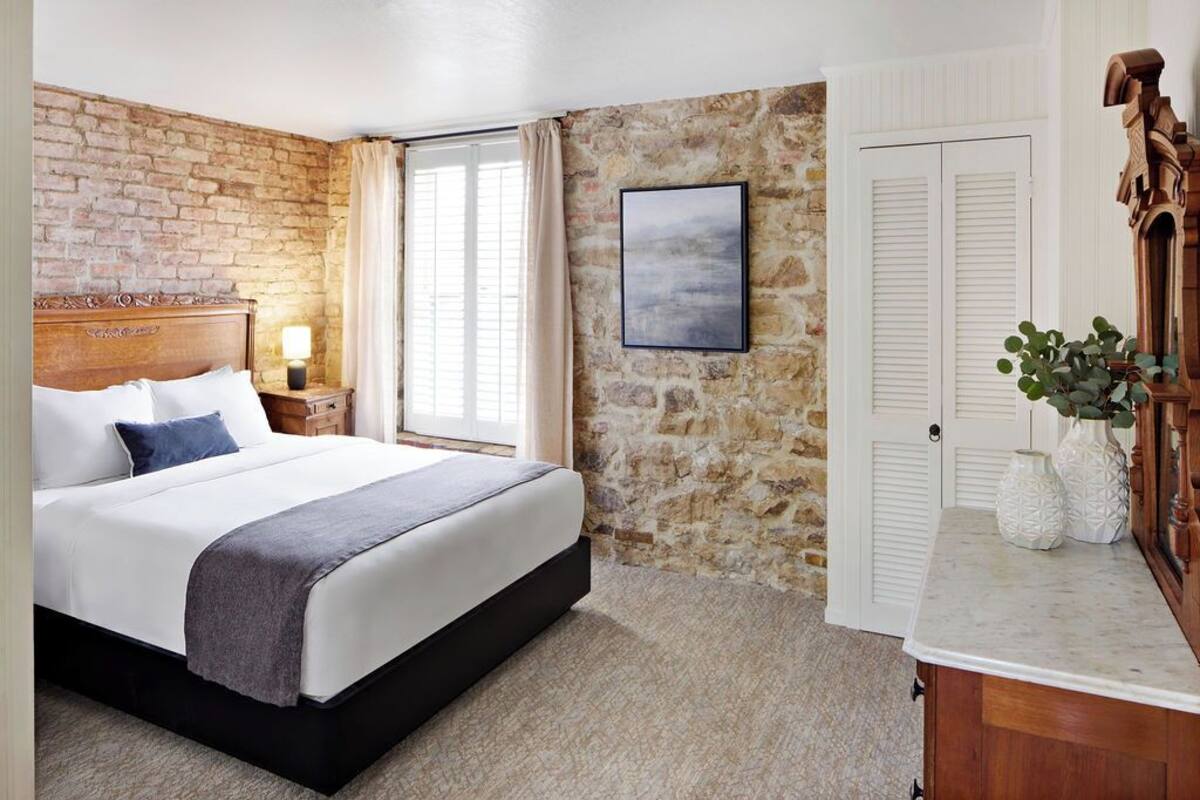 Un dormitorio con pared de piedra, una cama con ropa de cama blanca, una mesita de noche con lámpara, una ventana con cortinas y un aparador de madera con jarrones.