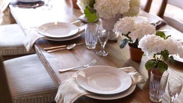 A wooden dining table set with plates, cutlery, glasses, and a potted plant with white flowers.