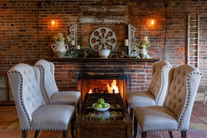 A fireplace with a brick wall, two upholstered chairs, a wooden coffee table with a bowl of apples, and decorative items on the mantel.