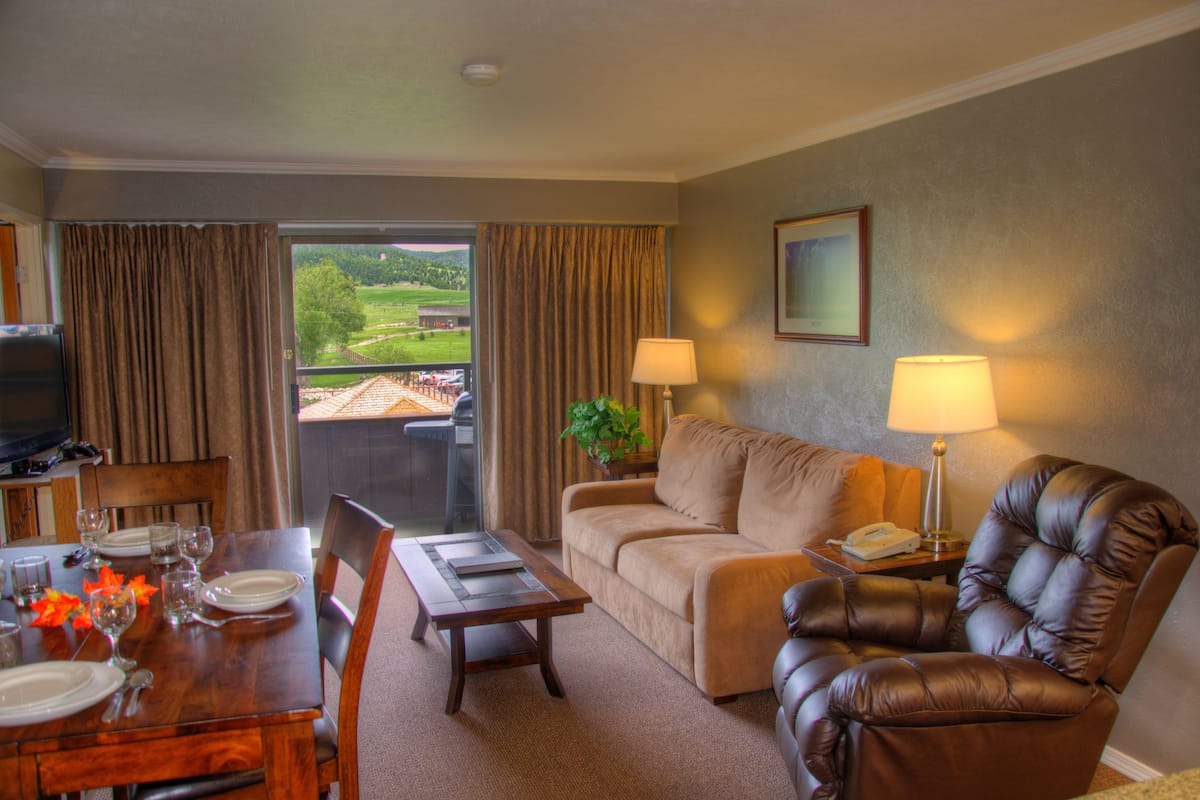 Ein Hotelzimmer mit Esstisch für vier Personen, einer Couch, einem Ledersessel, einem Fernseher und einem Balkon mit Vorhängen.