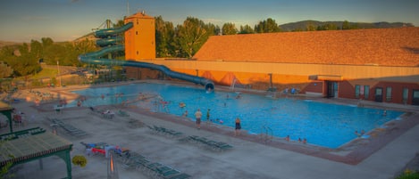 A swimming pool with a water slide, lounge chairs, and a covered area with tables and chairs.