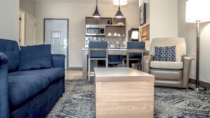 Ein modernes Wohnzimmer mit einem blauen Sofa, einem Holztisch, einem beigen Sessel und einer Stehlampe. Es gibt eine Küchenzeile mit einer Mikrowelle, einer Kaffeemaschine und einem Spülbecken.
