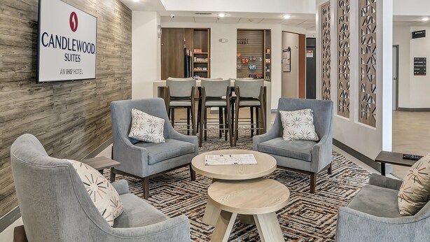 Eine moderne Hotelhalle mit grauen Sesseln, einem Holztisch und einer Rezeption. Ein Schild an der Wand zeigt 
