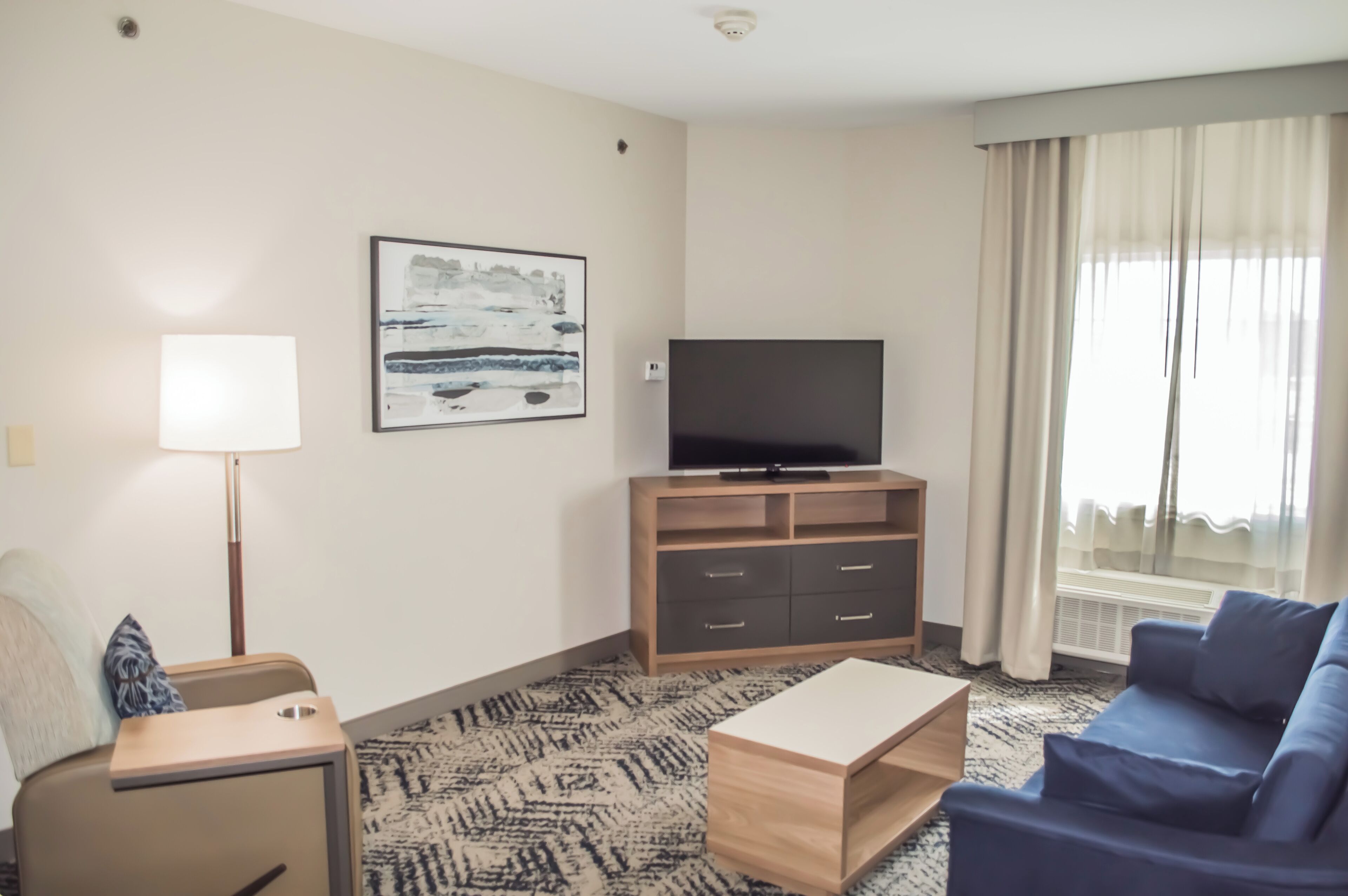 A hotel room with a flat-screen TV on a wooden stand, a sofa, a small table, a lamp, and a painting on the wall.