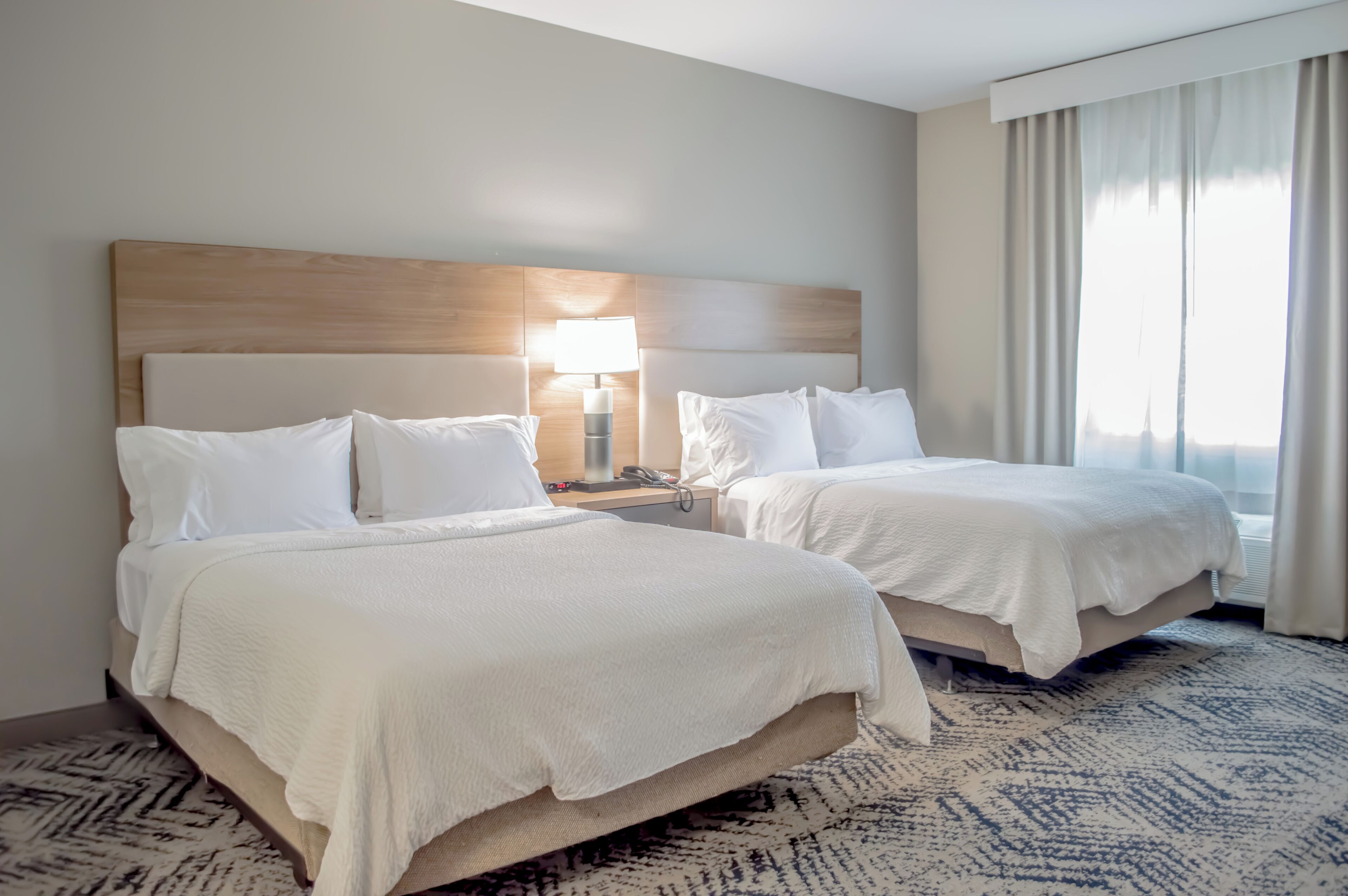 A hotel room with two beds, a bedside table with a lamp, and a large window with curtains.