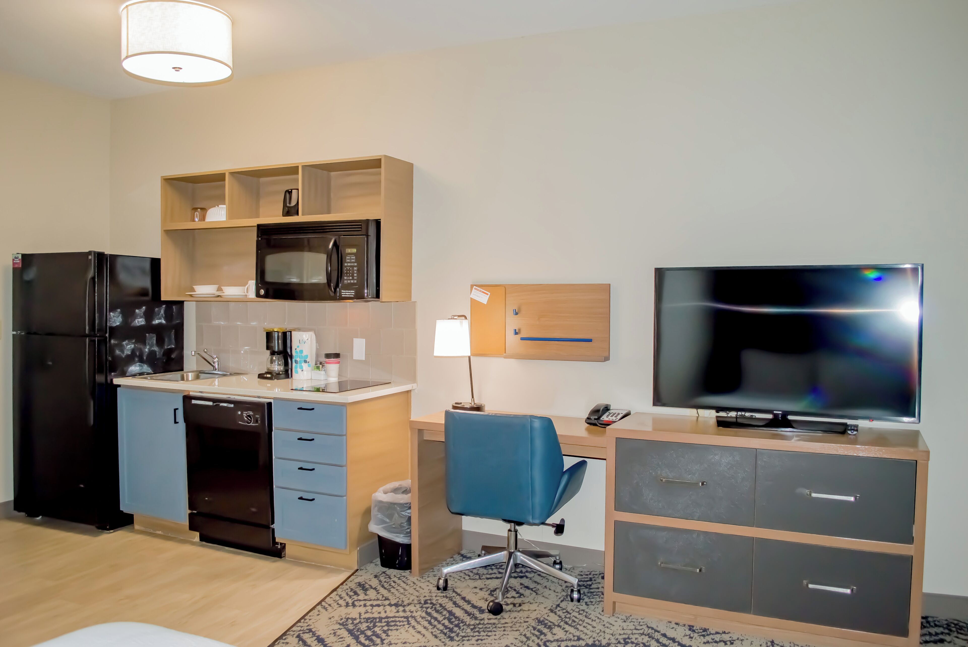 A compact hotel room with a kitchenette, a blue office chair, a flat-screen TV, and a wooden cabinet.