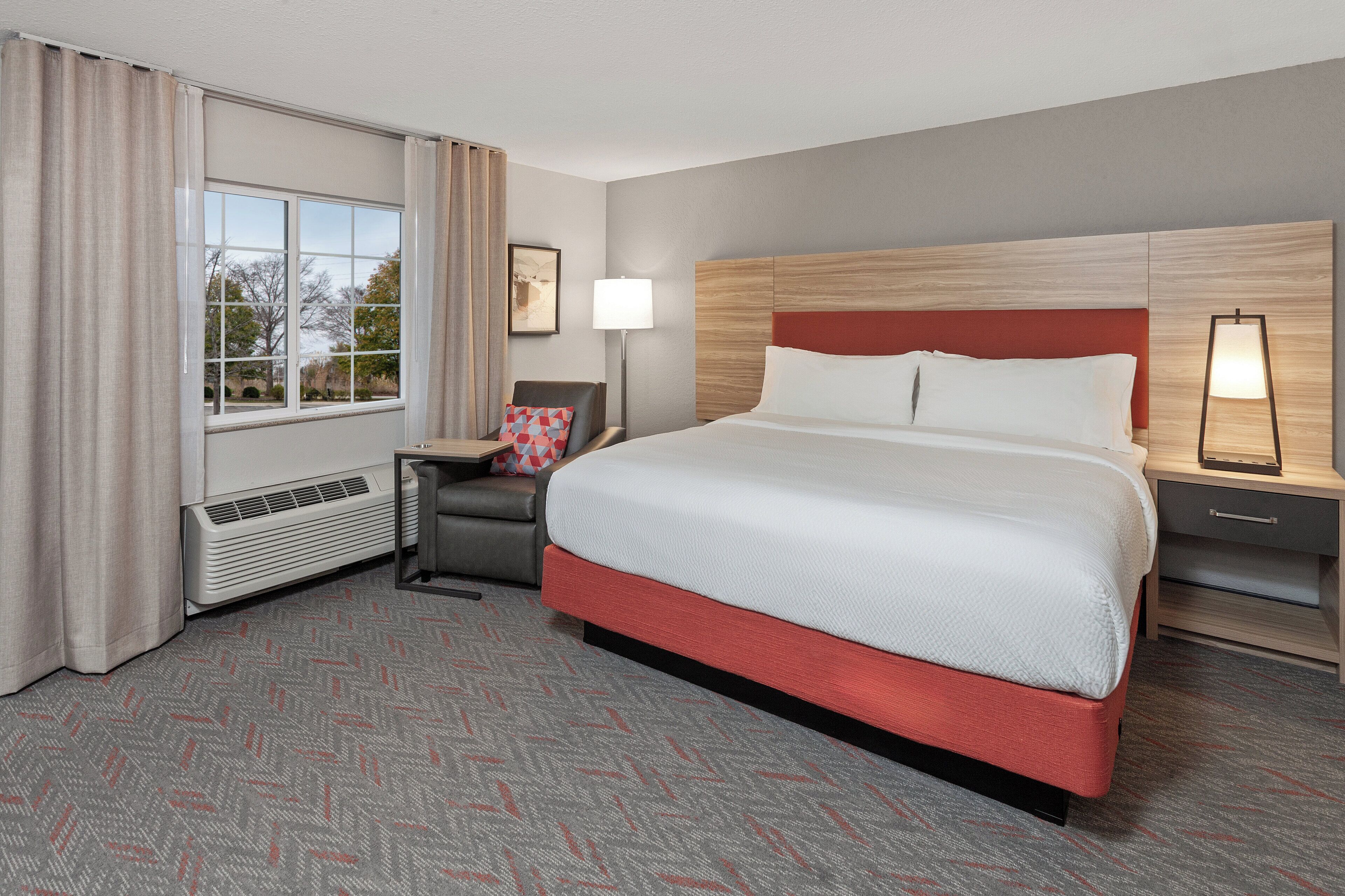 A hotel room with a large bed, a chair, a desk, a lamp, and a window with curtains.