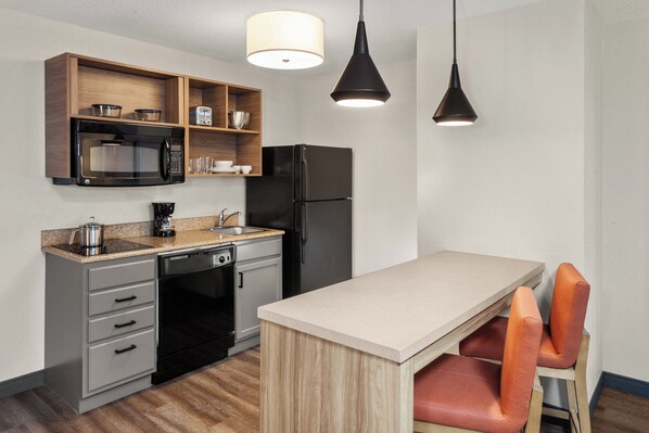 A compact kitchen with a microwave, refrigerator, and wooden shelves. There are two pendant lights above the counter. - Candlewood Suites Elgin by IHG (Elgin)