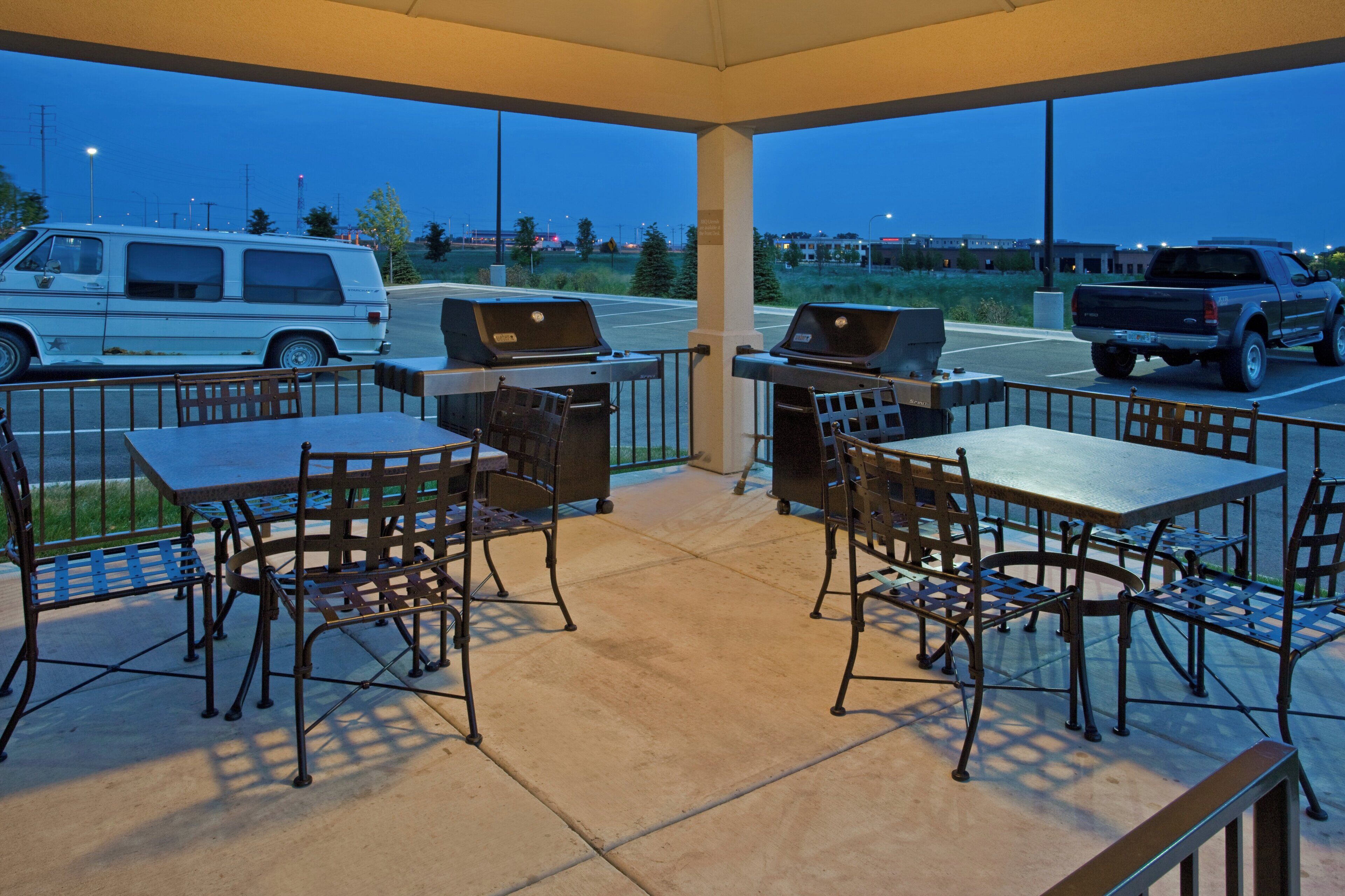 Un área de patio al aire libre con mesas y sillas, una parrilla y vistas a vehículos estacionados y edificios al fondo.