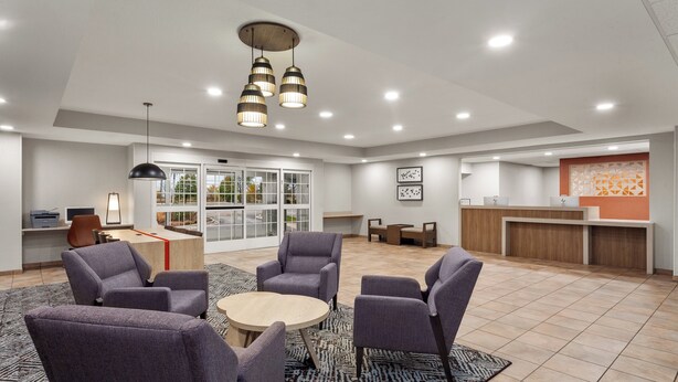 A spacious lobby with a reception desk, seating area with chairs and a round table, and a large window with a view of the outdoors.