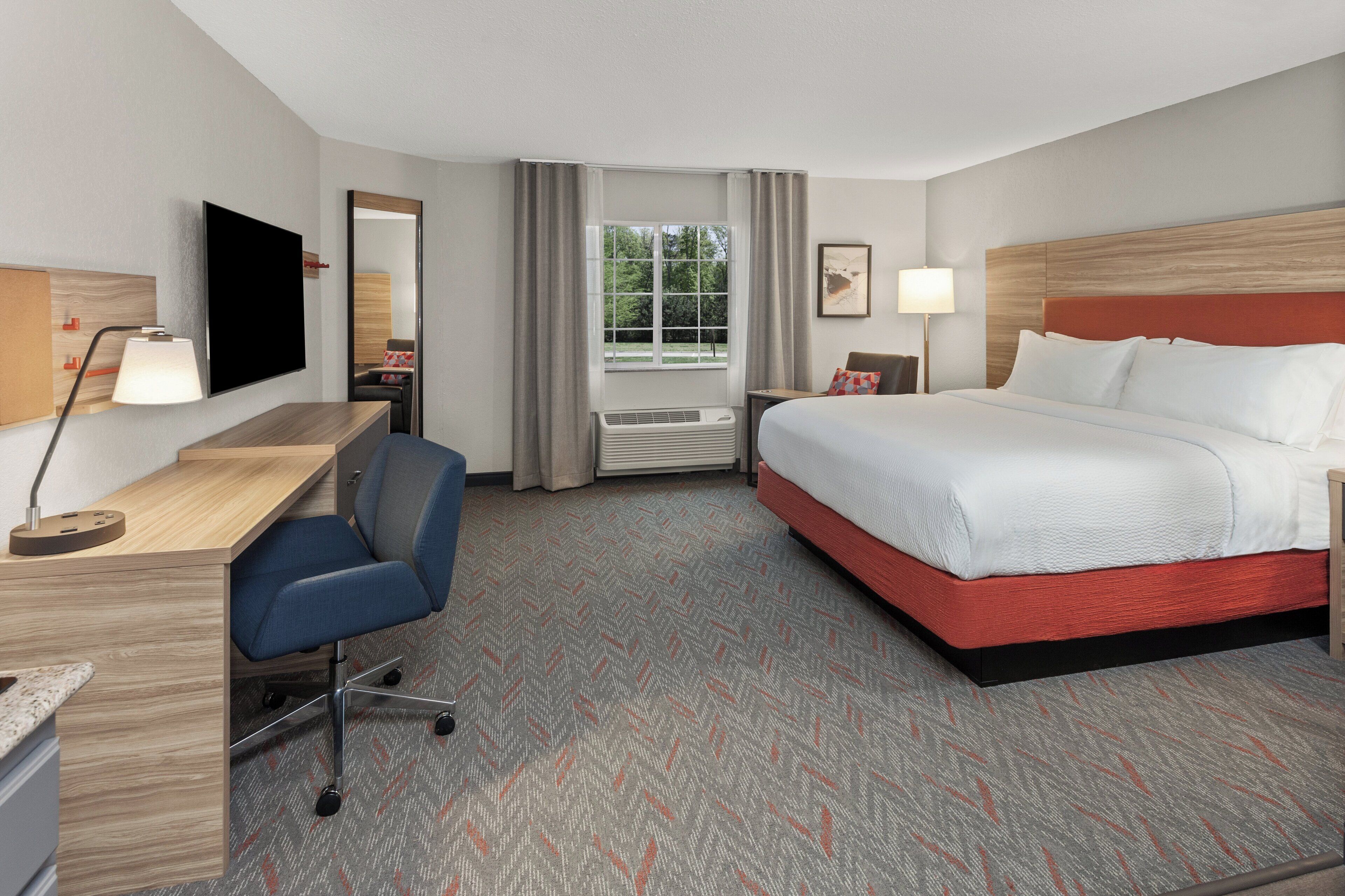 A hotel room with a bed, desk, chair, TV, window, and artwork.