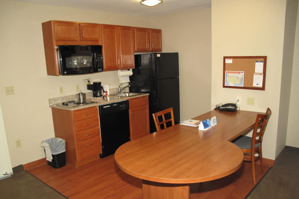 A compact kitchen with wooden cabinets, a black refrigerator, a microwave, and a dishwasher. There's a dining table with chairs and a bulletin board on the wall.