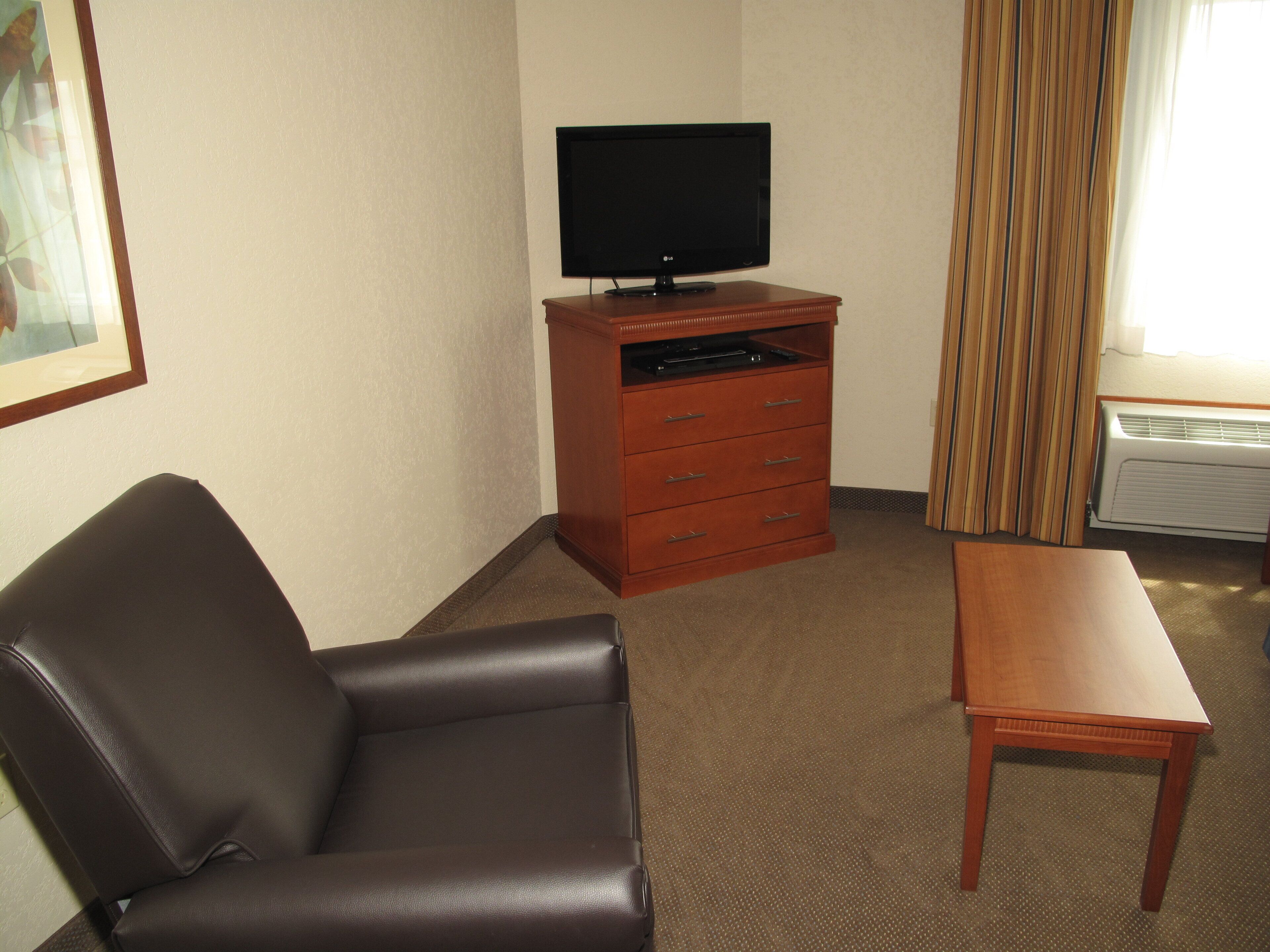 Une chambre d’hôtel avec un fauteuil en cuir, une table en bois, un téléviseur posé sur un meuble en bois et une fenêtre avec des rideaux.