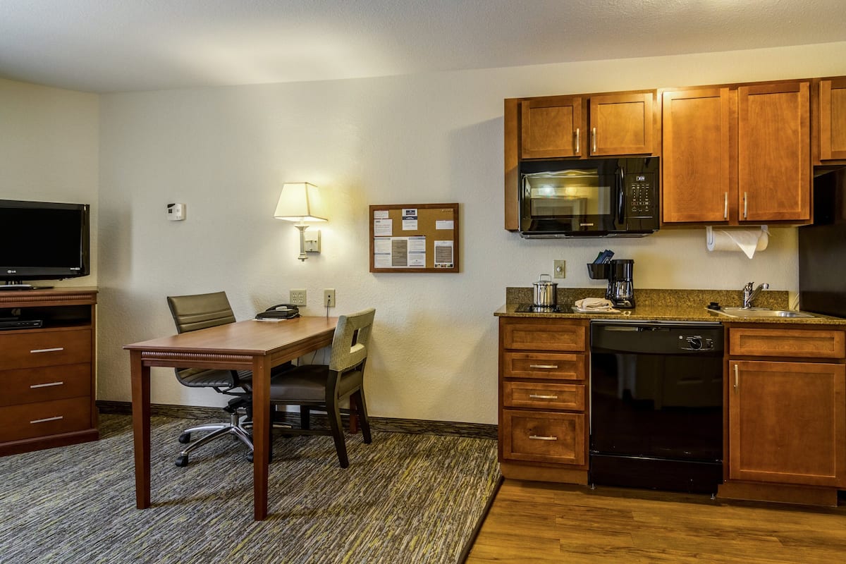 Ein kleines Hotelzimmer mit Schreibtisch aus Holz, Fernseher, Mikrowelle, Kaffeemaschine und Geschirrspüler.