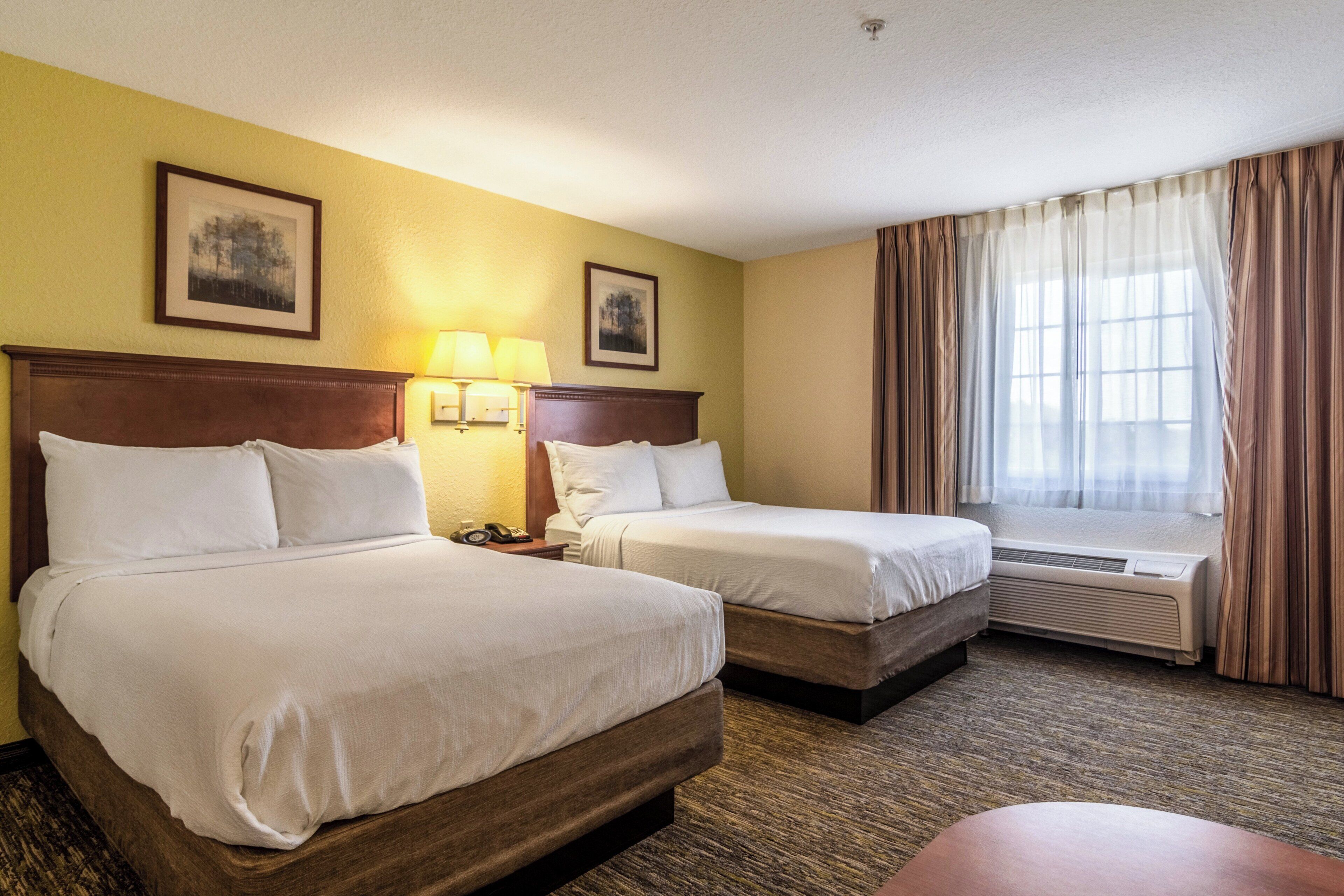 Une chambre d’hôtel avec deux lits, une fenêtre avec des rideaux, un climatiseur fixé au mur et deux tableaux encadrés.