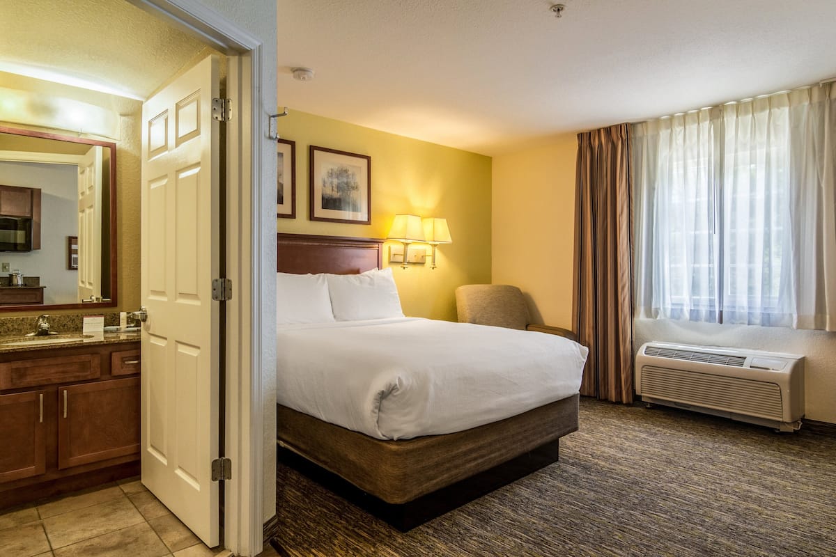 Ein Hotelzimmer mit Bett, Sessel, Schreibtisch mit Lampe, Fenster mit Vorhängen und einer Klimaanlage.