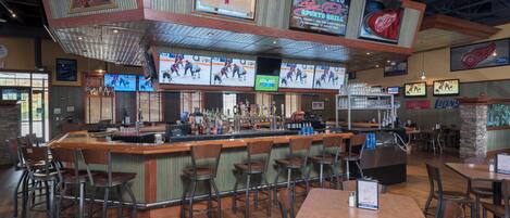 A sports bar with multiple screens displaying hockey games, a bar area with stools, and a seating section with tables and chairs.