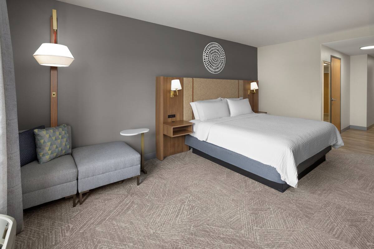 A hotel room with a bed, a sofa, a small table, a wall-mounted lamp, and a door.
