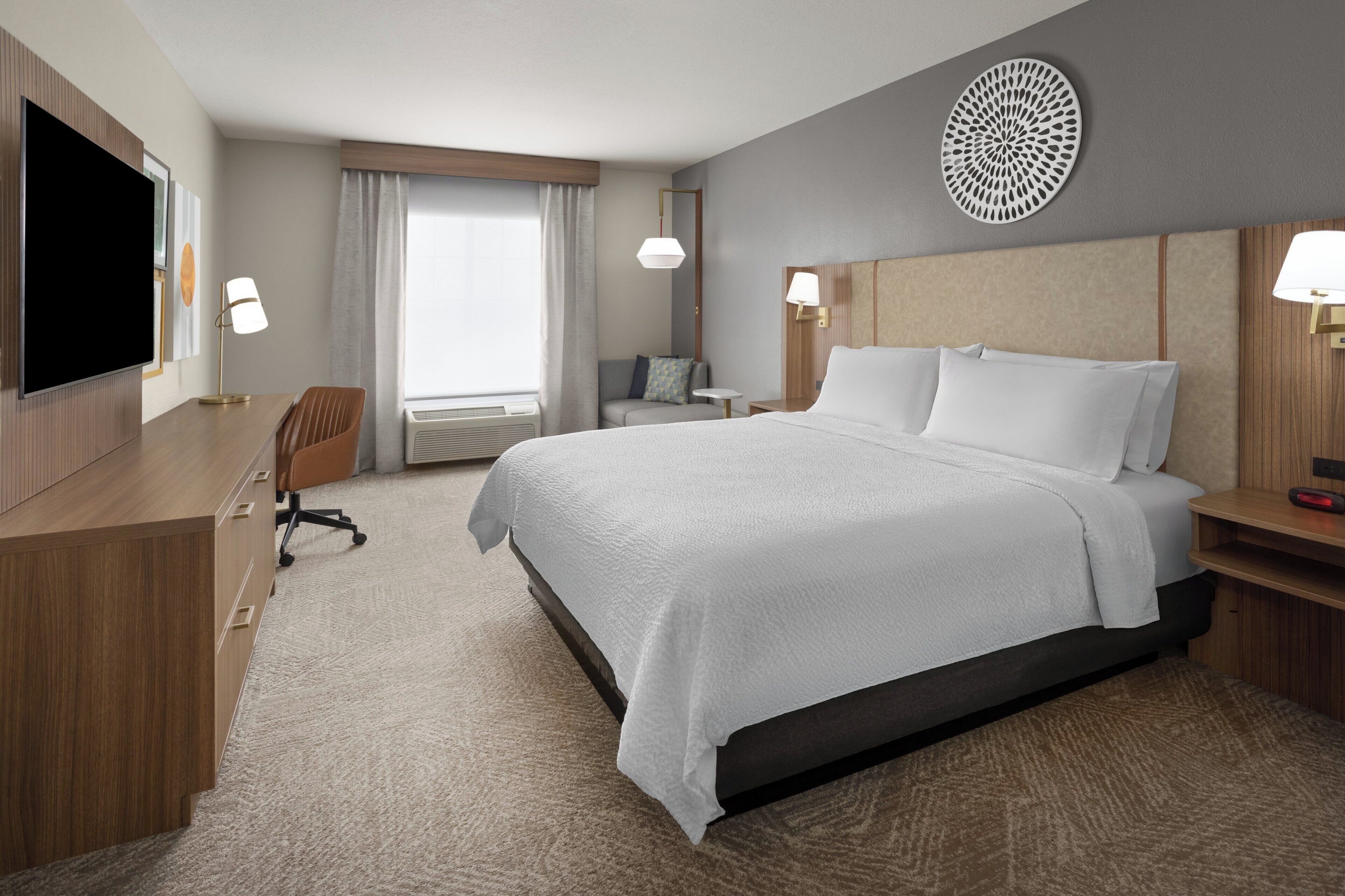 A hotel room with a large bed, a desk with a chair, a TV, and a round decorative wall piece.