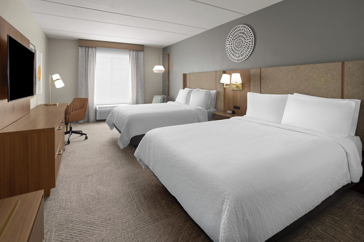 A hotel room with two beds, a desk, a TV, and a window with curtains.