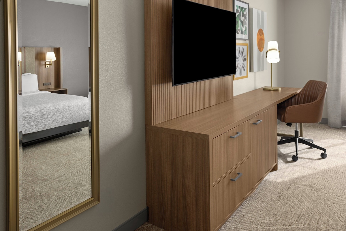 A hotel room with a bed, a desk with a chair, a television, and a mirror.