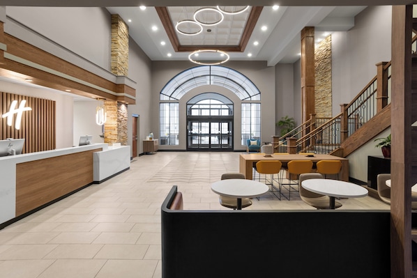 A spacious lobby with a reception desk, a staircase, and a seating area with tables and chairs.