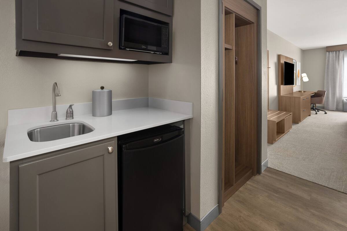 A compact kitchen with a sink, microwave, and dishwasher, leading to a bedroom with a desk and chair.