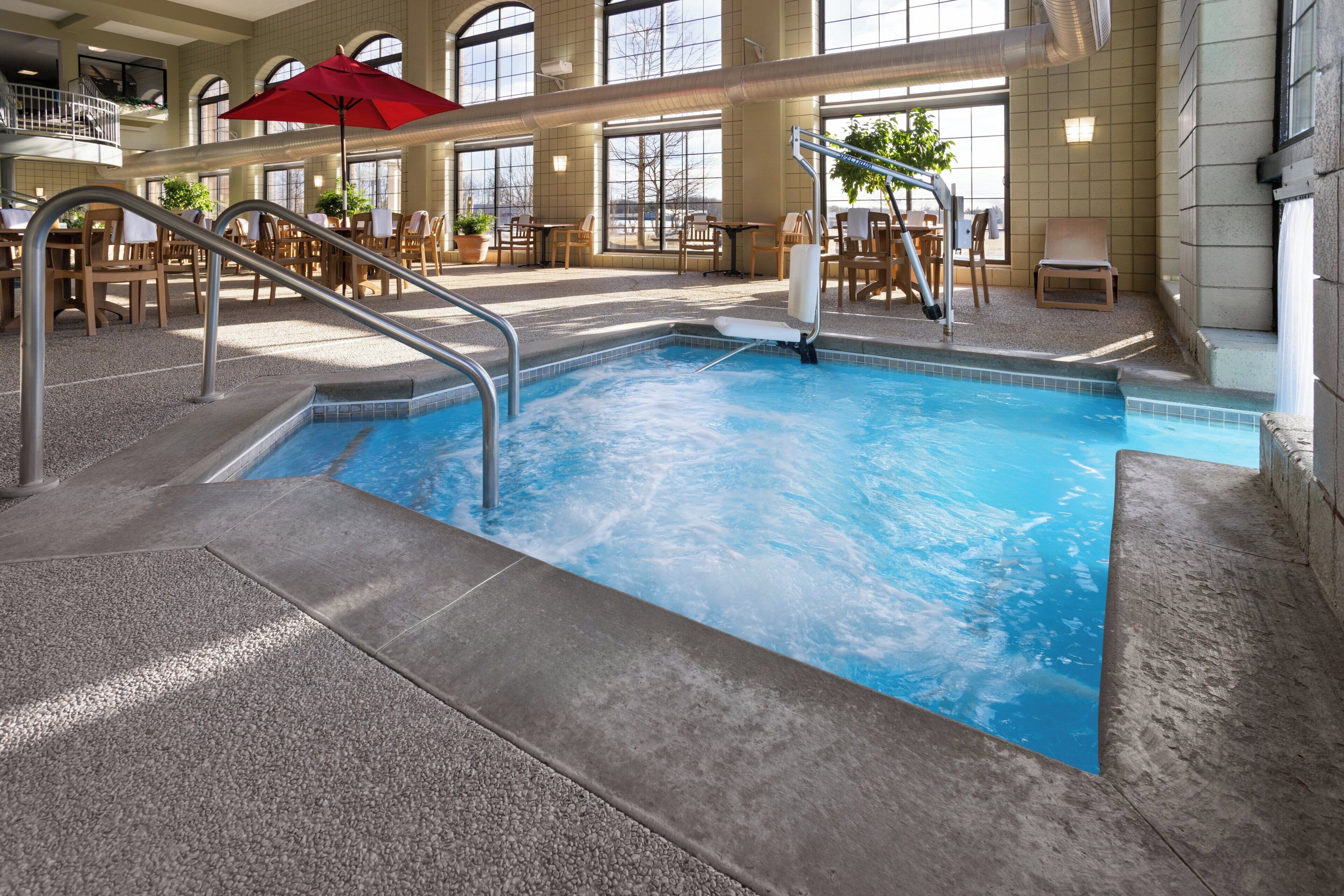 An indoor swimming pool with a red umbrella, lounge chairs, and a view of the outdoors through large windows.