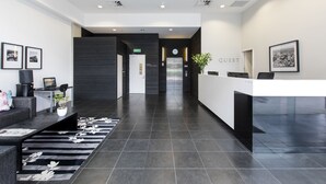 A modern hotel lobby with a reception desk, a seating area, and a staircase leading to an elevator. - Quest Carlaw Park (Auckland)