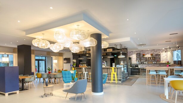 A modern hotel lobby with a central seating area, a bar, and a kitchen visible in the background. - Hôtel ibis Lyon Sud Vienne Saint Louis (Vienne)