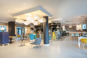 Een moderne hotel lobby met een centraal zitgedeelte, een bar en een keuken in de achtergrond.