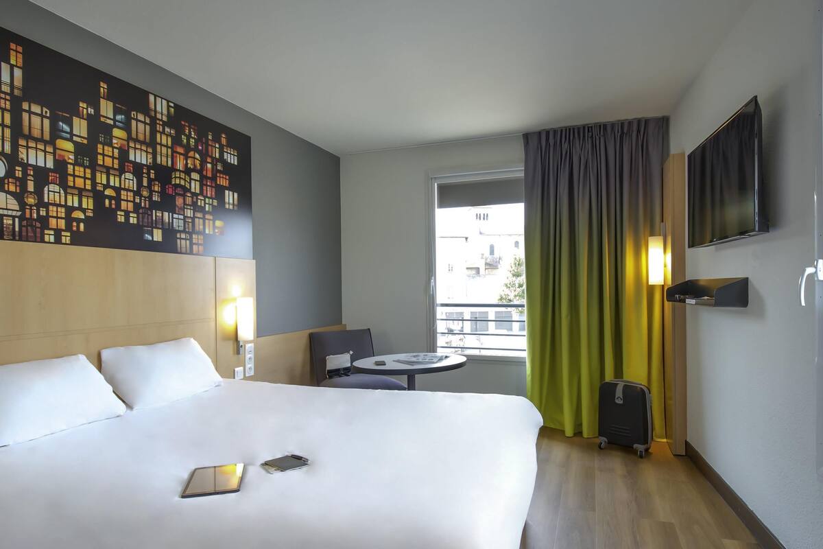 Een hotelkamer met een groot bed, een klein tafeltje, een stoel, een televisie en een raam met gordijnen.