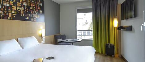 Habitación de hotel con una cama grande, una mesita, una silla, un televisor y una ventana con cortinas.