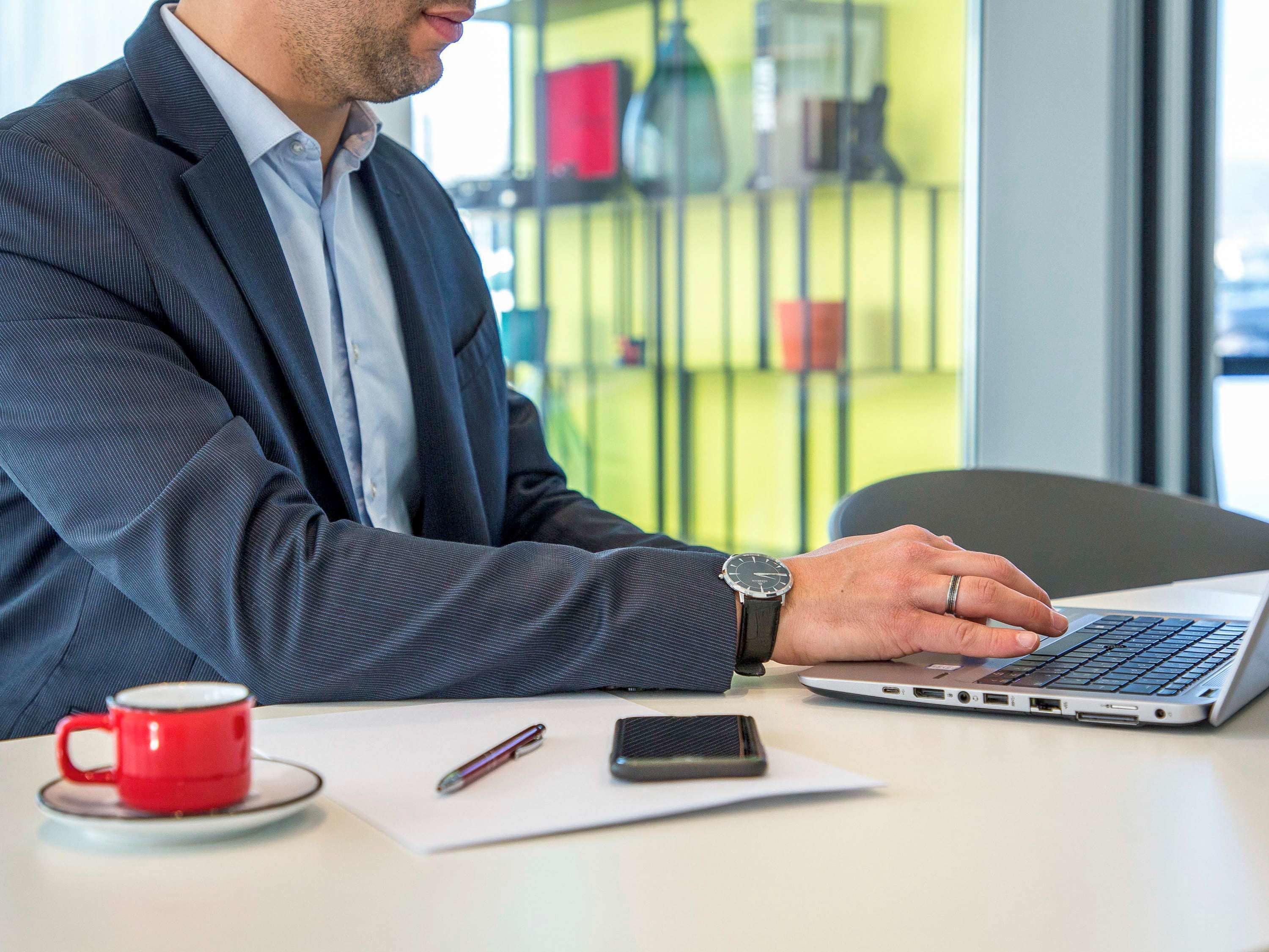 A man in a suit working on a laptop at a table with a red mug, a pen, and a smartphone.