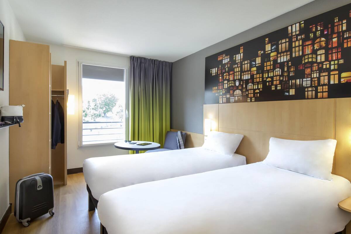 Een hotelkamer met twee bedden, een kleine tafel, een stoel, een koffer en een groot raam met gordijnen.