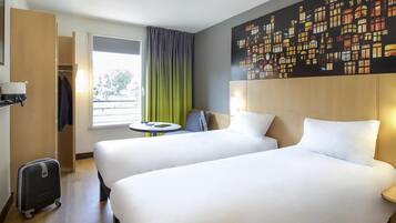 A hotel room with two beds, a small table, a chair, a suitcase, and a large window with curtains.