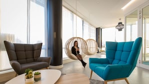 A modern office space with comfortable seating, including a blue armchair and a grey sofa, near large windows with natural light. - Hôtel ibis Lyon Sud Vienne Saint Louis (Vienne)