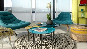 A modern living room with teal armchairs, a glass coffee table, a patterned rug, and a view through large windows. - Hôtel ibis Lyon Sud Vienne Saint Louis (Vienne)