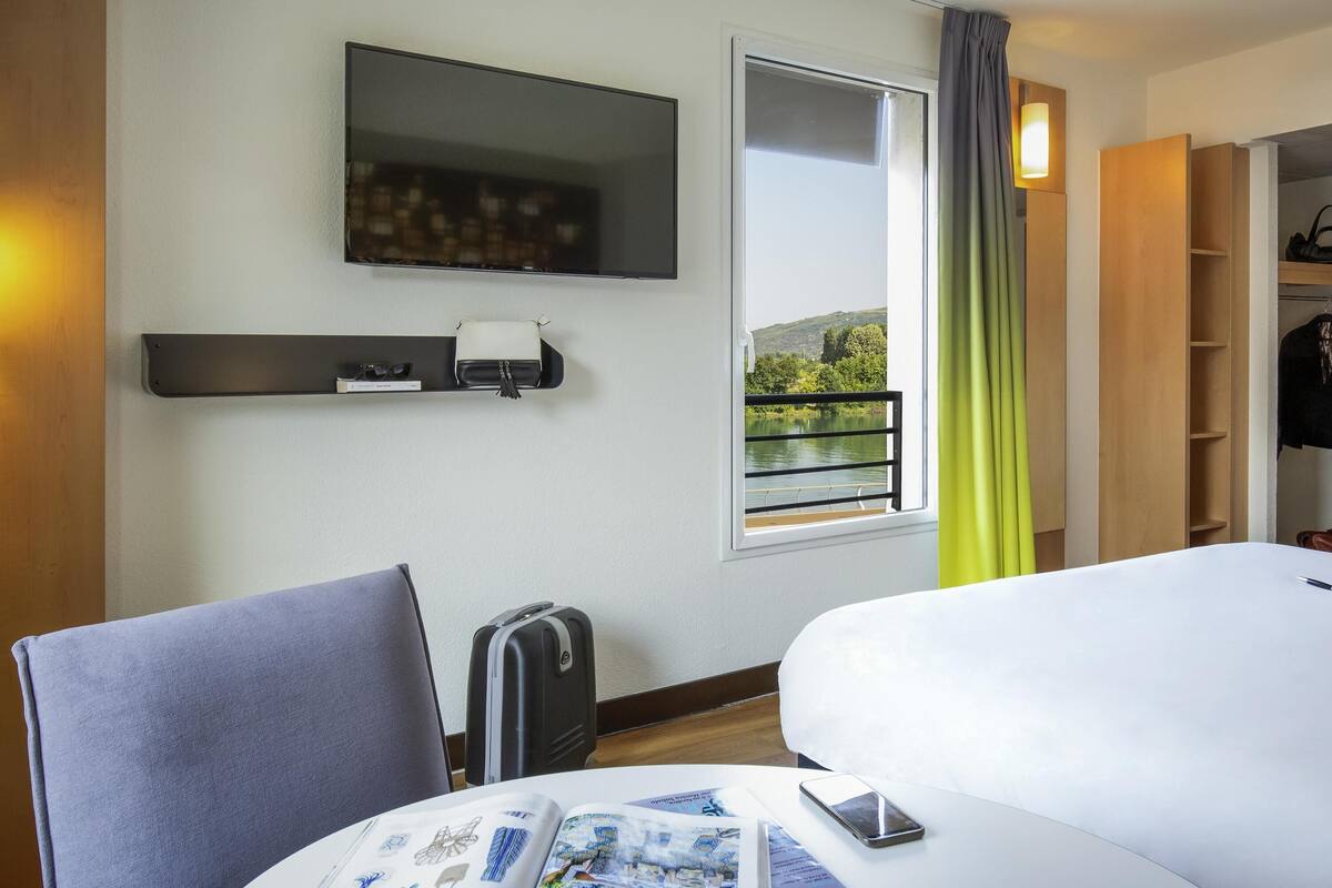 Une chambre d’hôtel comprenant un lit, un bureau avec une chaise, un téléviseur fixé au mur, une fenêtre donnant sur un lac et une petite étagère avec une lampe.