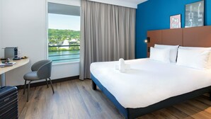 A hotel room with a bed, a desk, a chair, a suitcase, and a view of a bridge and greenery. - Hôtel ibis Lyon Sud Vienne Saint Louis (Vienne)